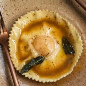 An egg yolk raviolo on a plate topped with brown butter sauce and crispy sage.