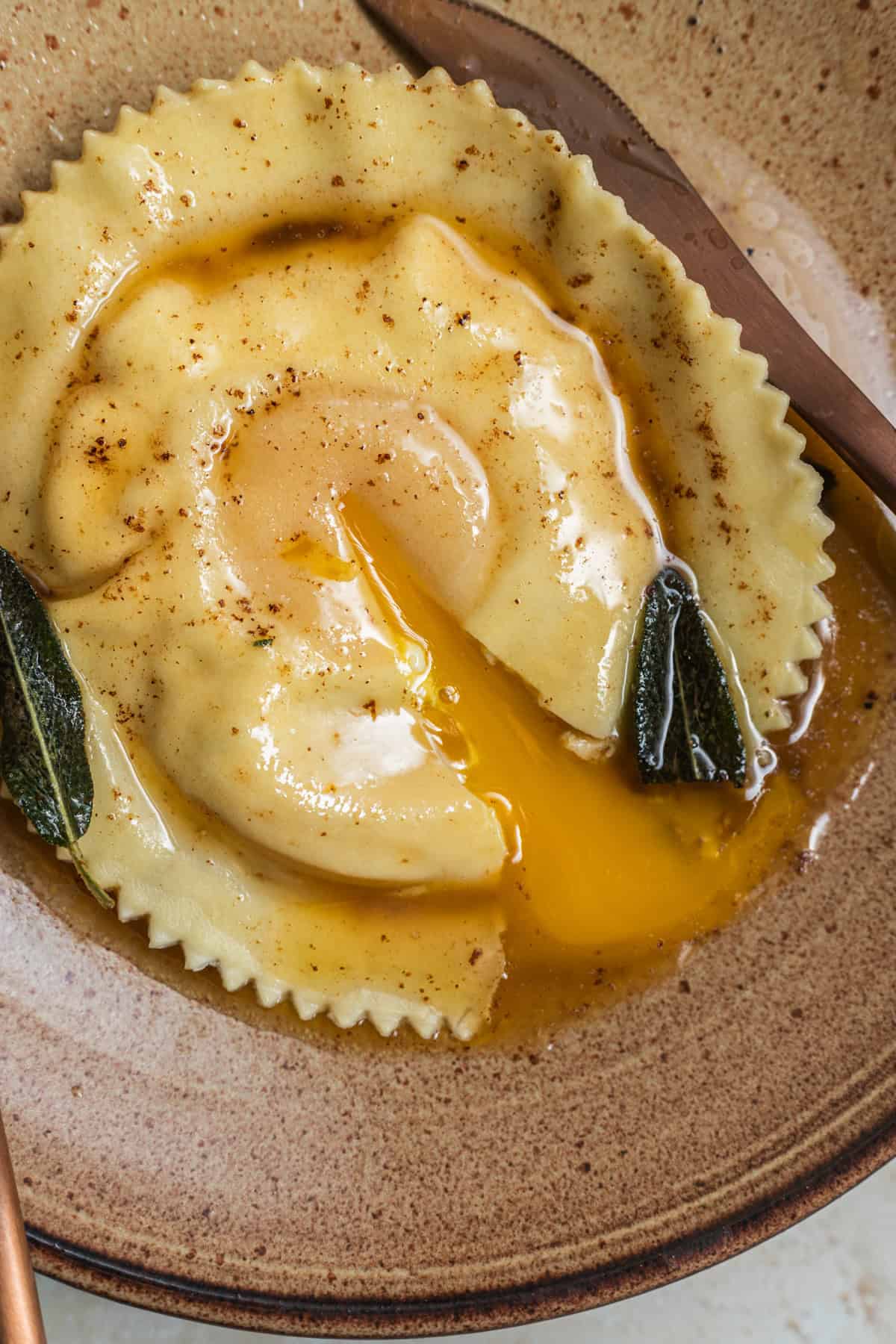 An egg yolk raviolo split open with the yolk oozing out in a pool of brown butter and sage leaves.