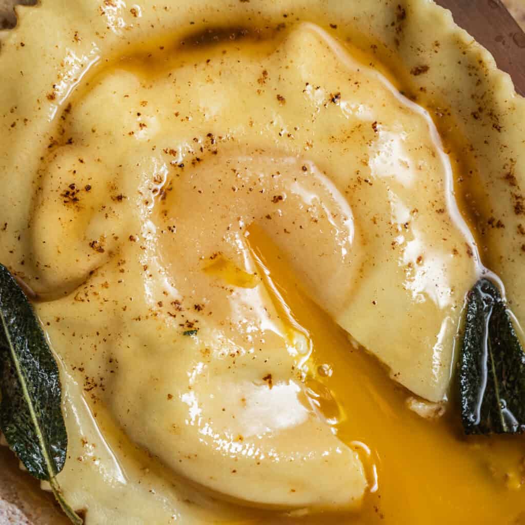 An egg yolk streaming out of a raviolo in a brown butter sauce with crispy sage.