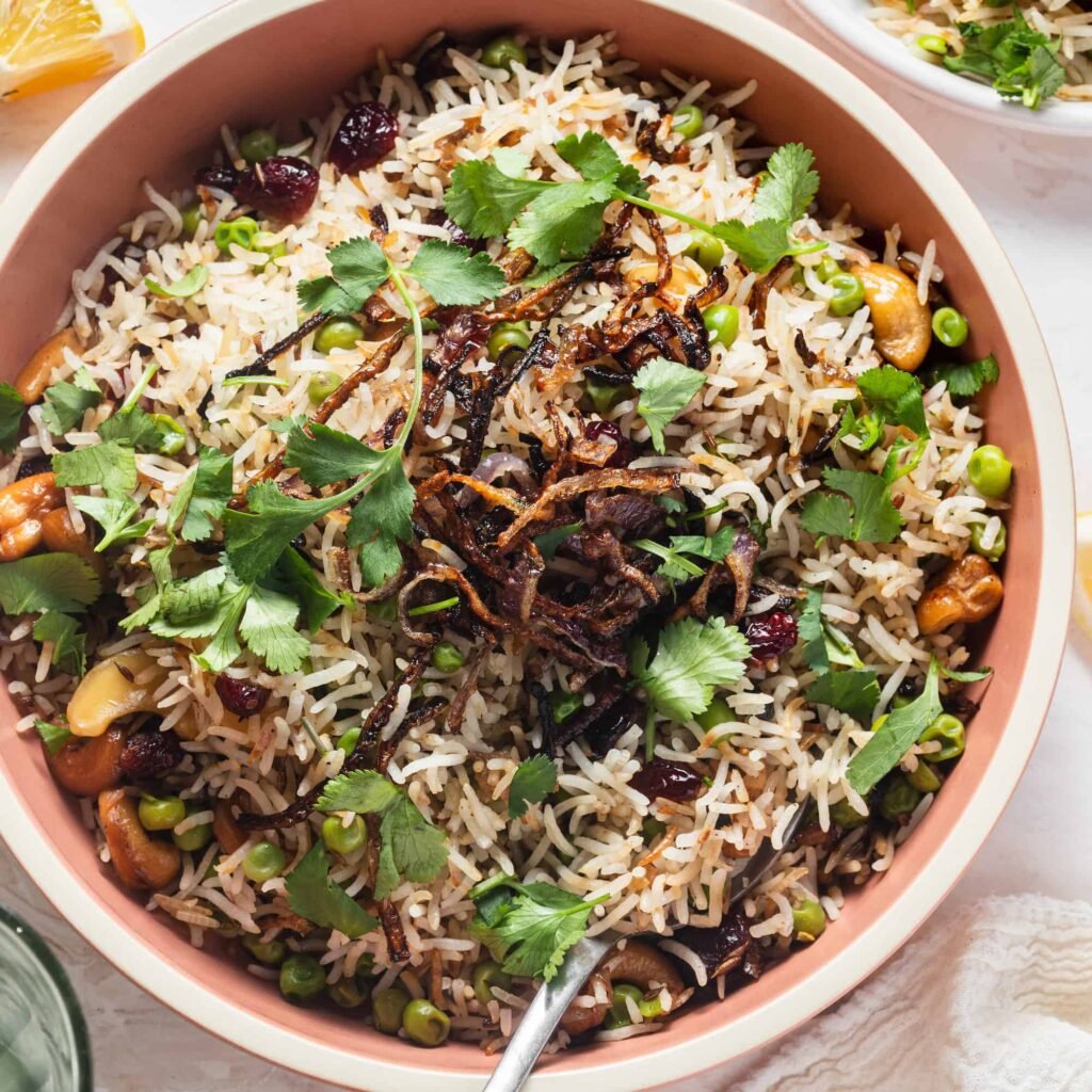 A bowl of pulao topped with peas, fresh cilantro, crispy fried onions, and cashews.