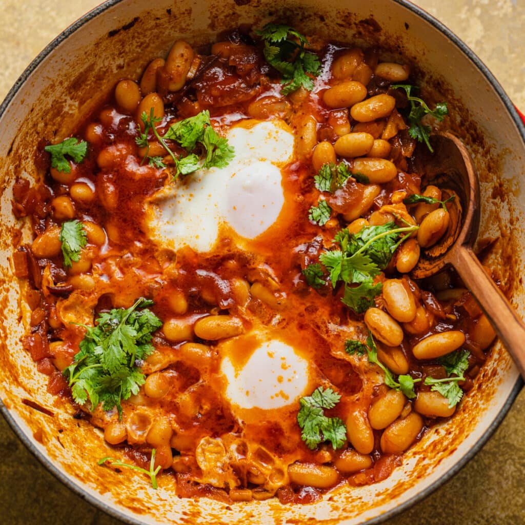 Skillet of beans and poached eggs in a tomato sauce with a wooden spoon.