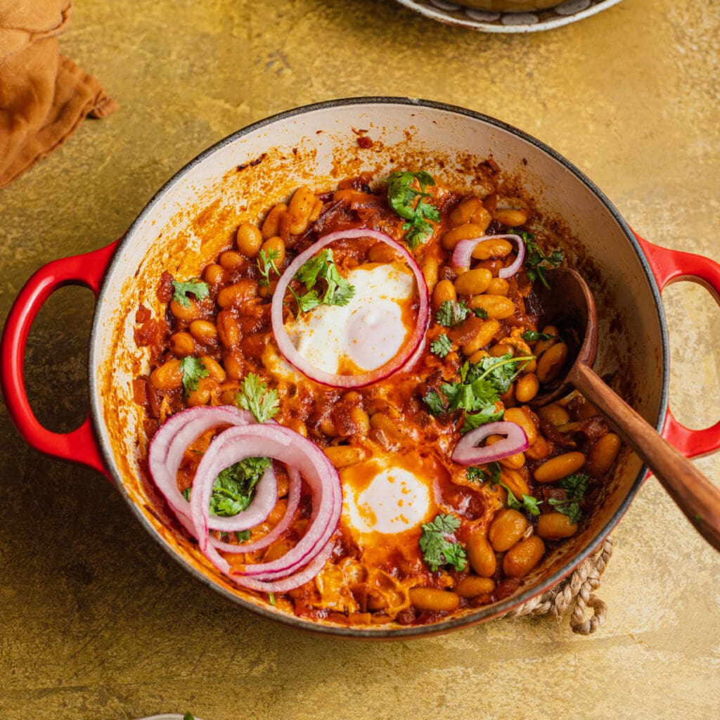 Skillet of beans and poached eggs in a tomato sauce topped with red onion and cilantro.