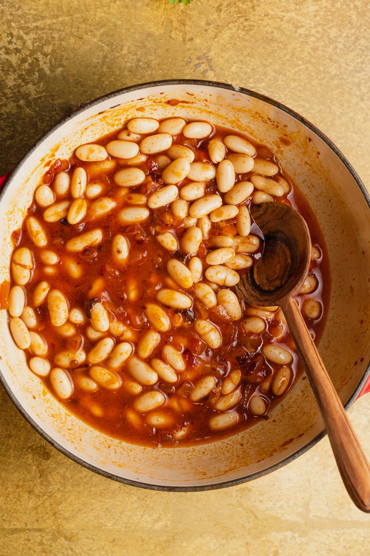 A skillet of white beans in a tomato-chipotle sauce with a wooden spoon.