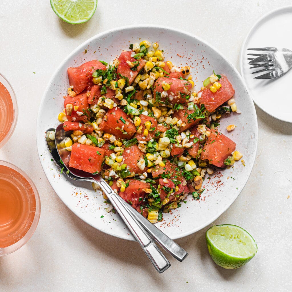 Bowl of watermelon jalapeno and corn salad with glasses and silverware in the background.
