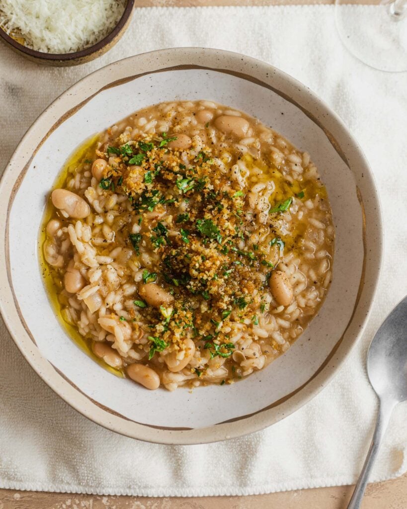 A bowl of risotto with cannellini beans and a breadcrumb topping on a white kitchen towel with a spoon next to it.