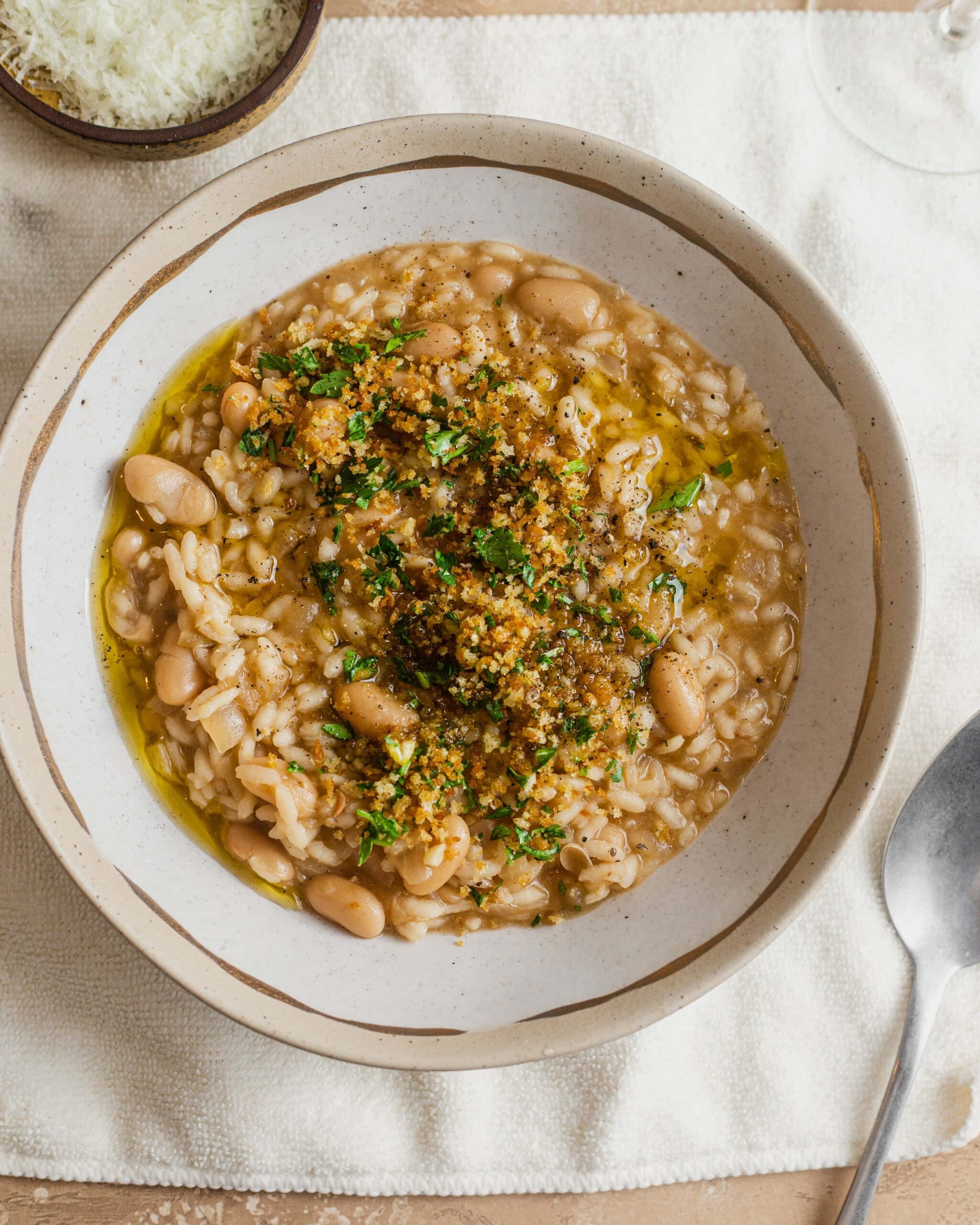 A bowl of risotto with cannellini beans and a breadcrumb topping on a white kitchen towel with a spoon next to it.