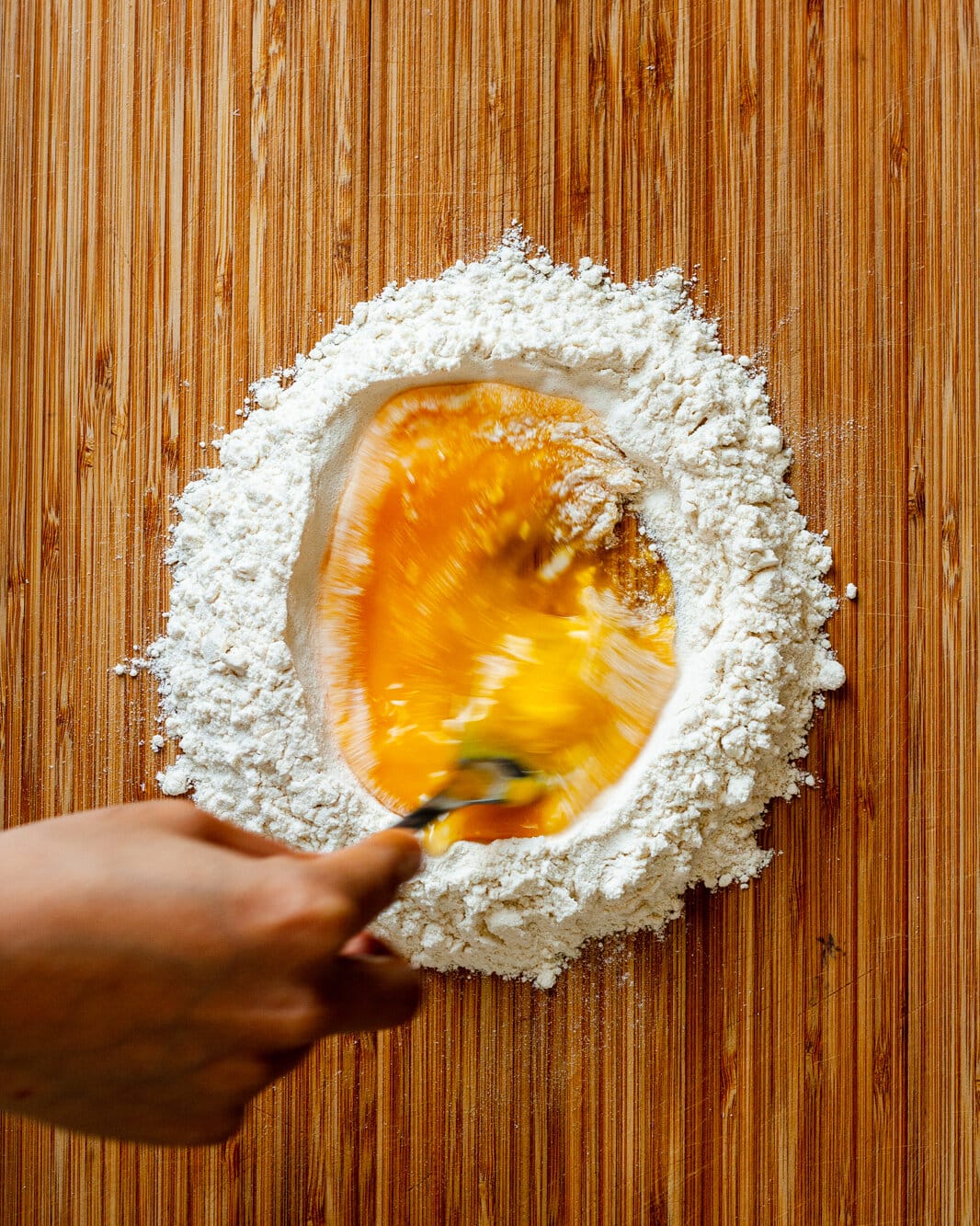 Hand whisking eggs into flour for pasta dough on a wooden cutting board.