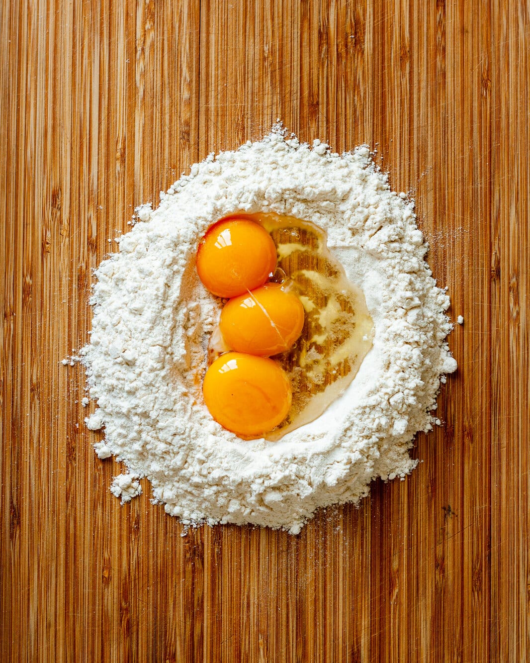 Well of flour filled with 3 eggs on a wooden cutting board.