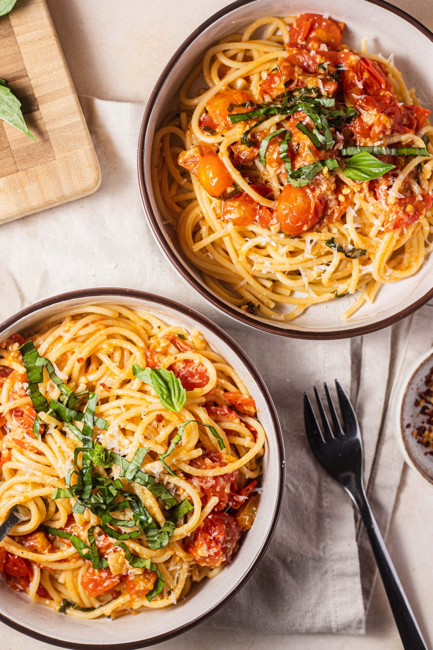 Two bowls of pasta with cherry tomatoes and basil and a cutting board in the background.