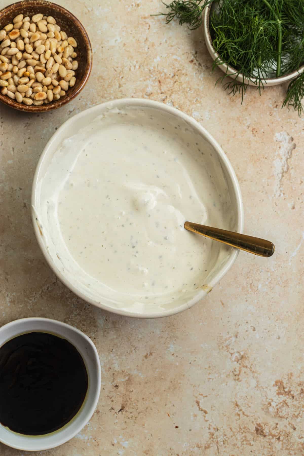 A bowl of yogurt with bowls of pine nuts, dill, and pomegranate molasses in the background.
