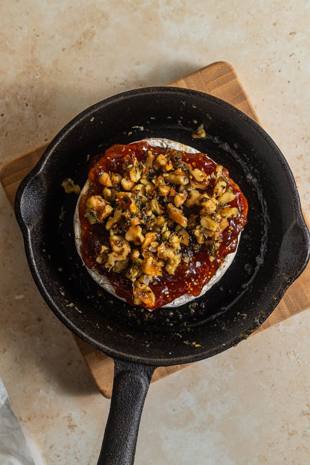 A cast-iron skillet set atop a wooden cutting board with a wheel of brie cheese topped with jam and nuts.