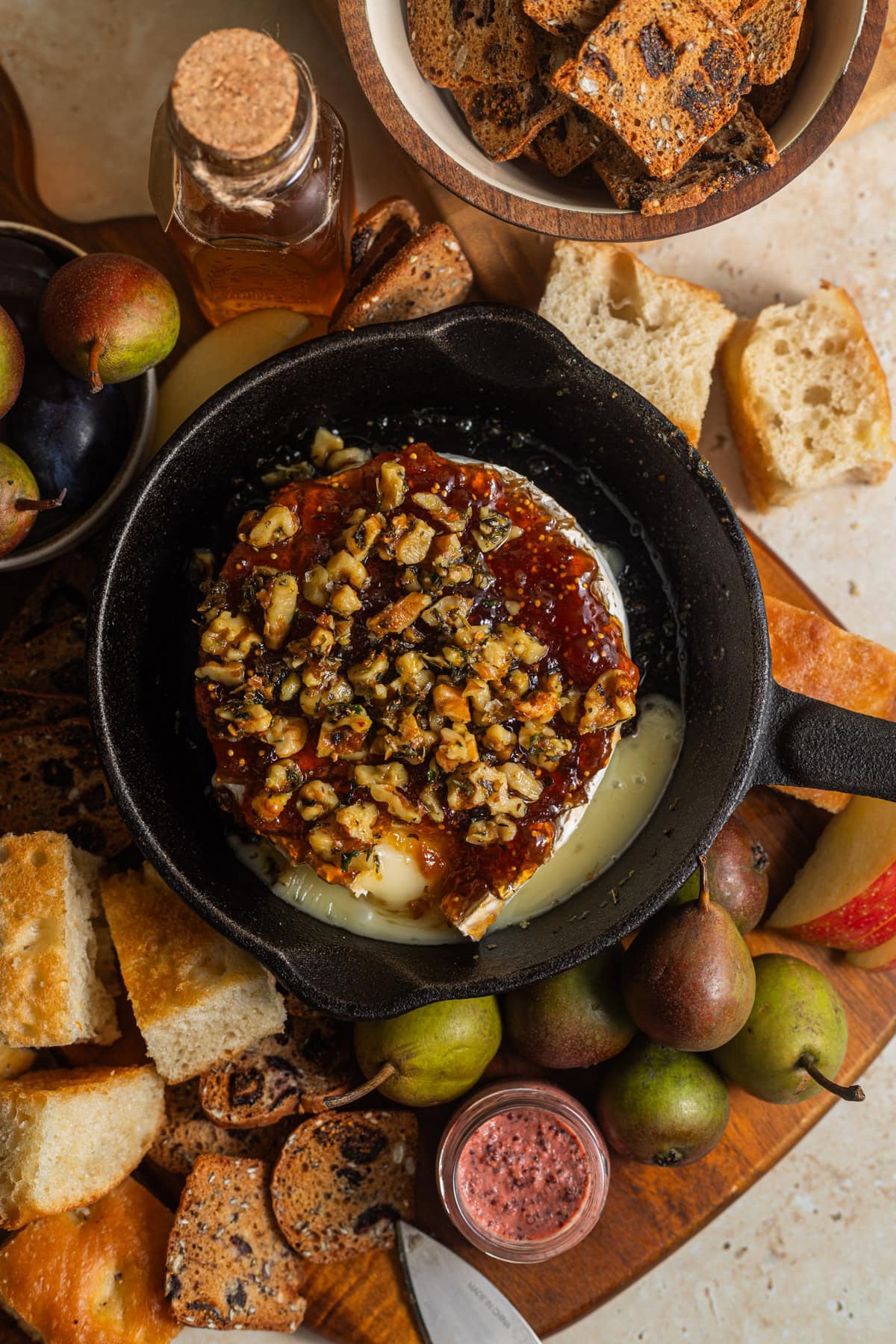 A wooden cheese board topped with pieces of focaccia, fresh pears, apples, and a skillet of baked brie.