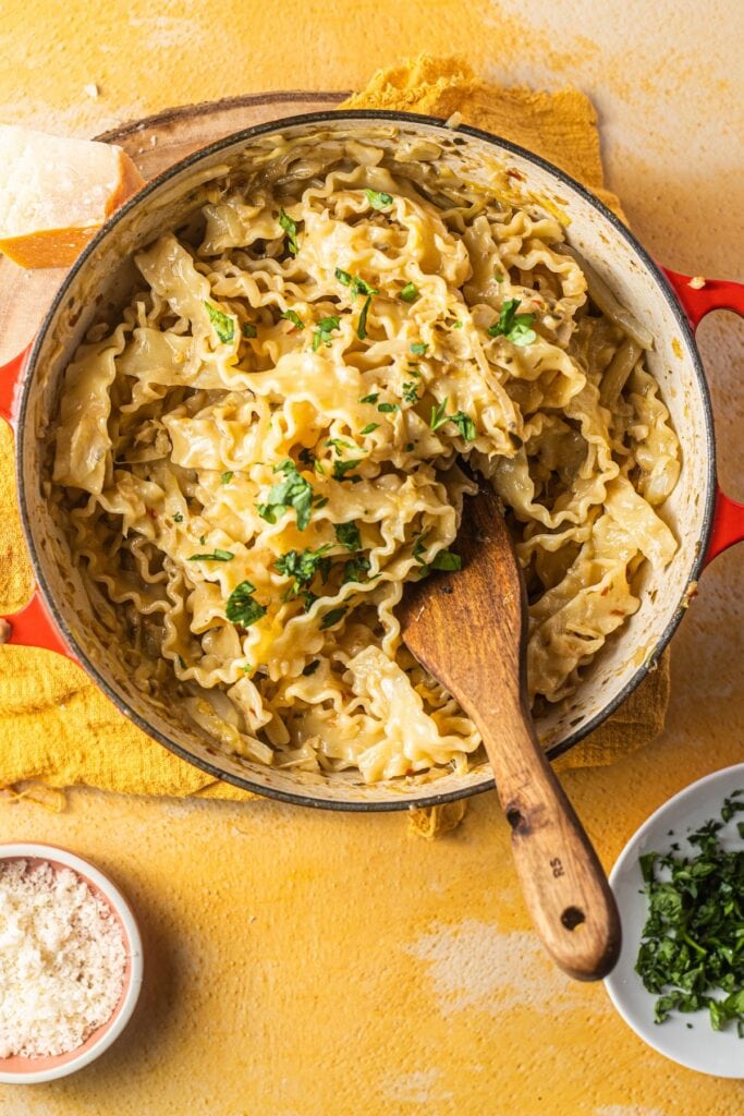 Mafaldine pasta in a dutch oven with bowls of cheese and herbs in the background.