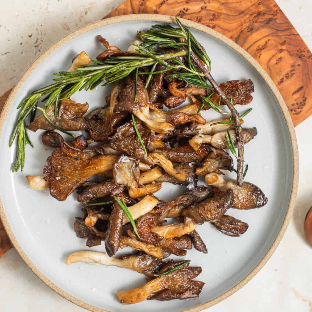 A plate of crispy sauteed blue oyster mushrooms with a sprig of rosemary on a wooden cutting board.