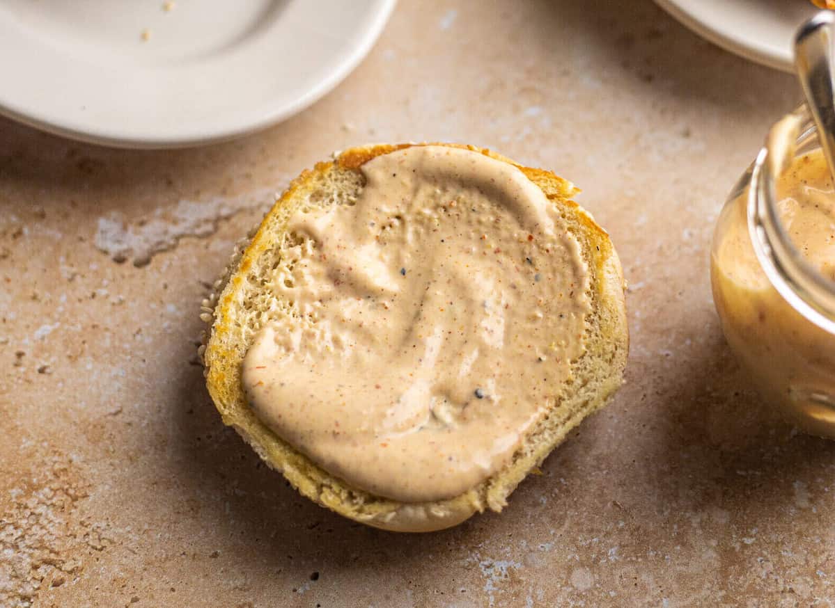 A burger bun with creamy sauce on top next to a jar of sauce.