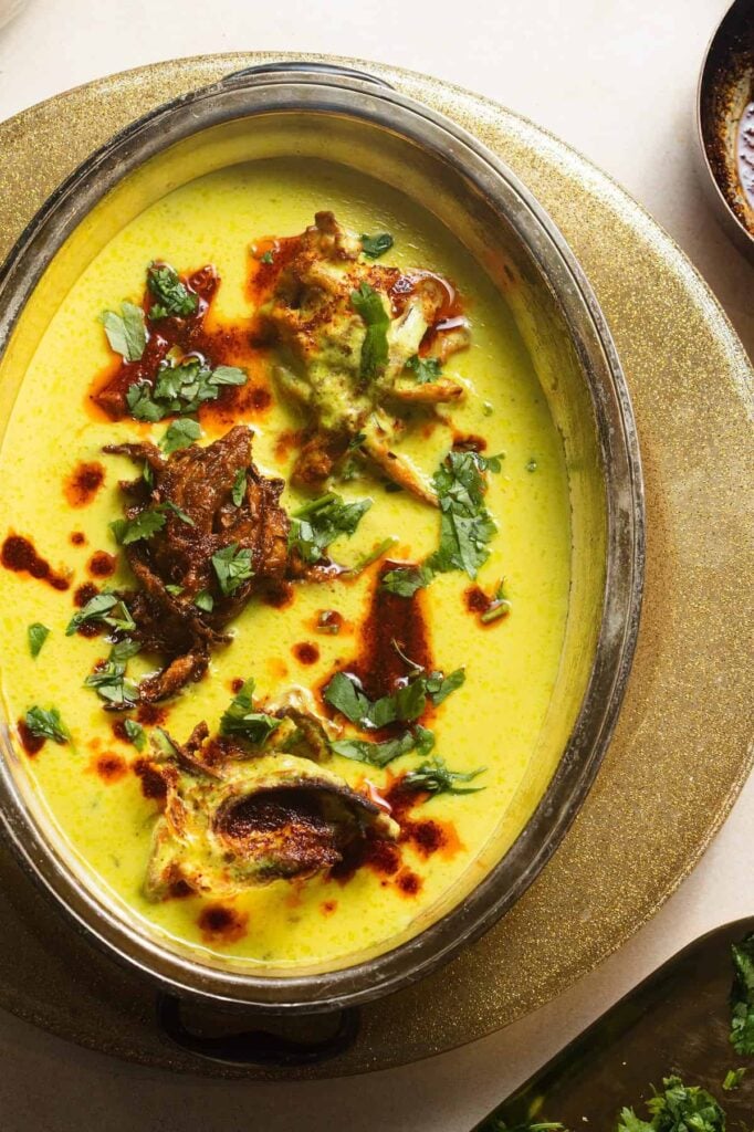 A metal bowl with kadhi topped with onion pakoras, chili oil, and fresh cilantro.