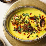 A metal bowl filled with a creamy yellow gravy topped with onion pakoras, chili oil, and fresh herbs set over a glittery plate.