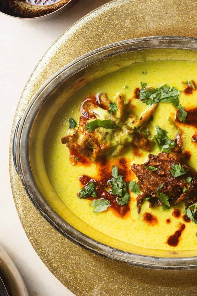 A metal bowl filled with a creamy yellow gravy topped with onion pakoras, chili oil, and fresh herbs set over a glittery plate.