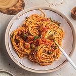 A bowl of spaghetti coated in a tomato sauce and topped with capers with a drink and bowl of cheese in the background.