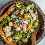 A wooden salad bowl filled with tuna Caesar salad and two wooden spoons ready for serving.