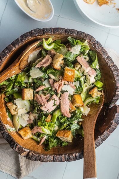 A wooden salad bowl filled with tuna Caesar salad and two wooden spoons ready for serving.