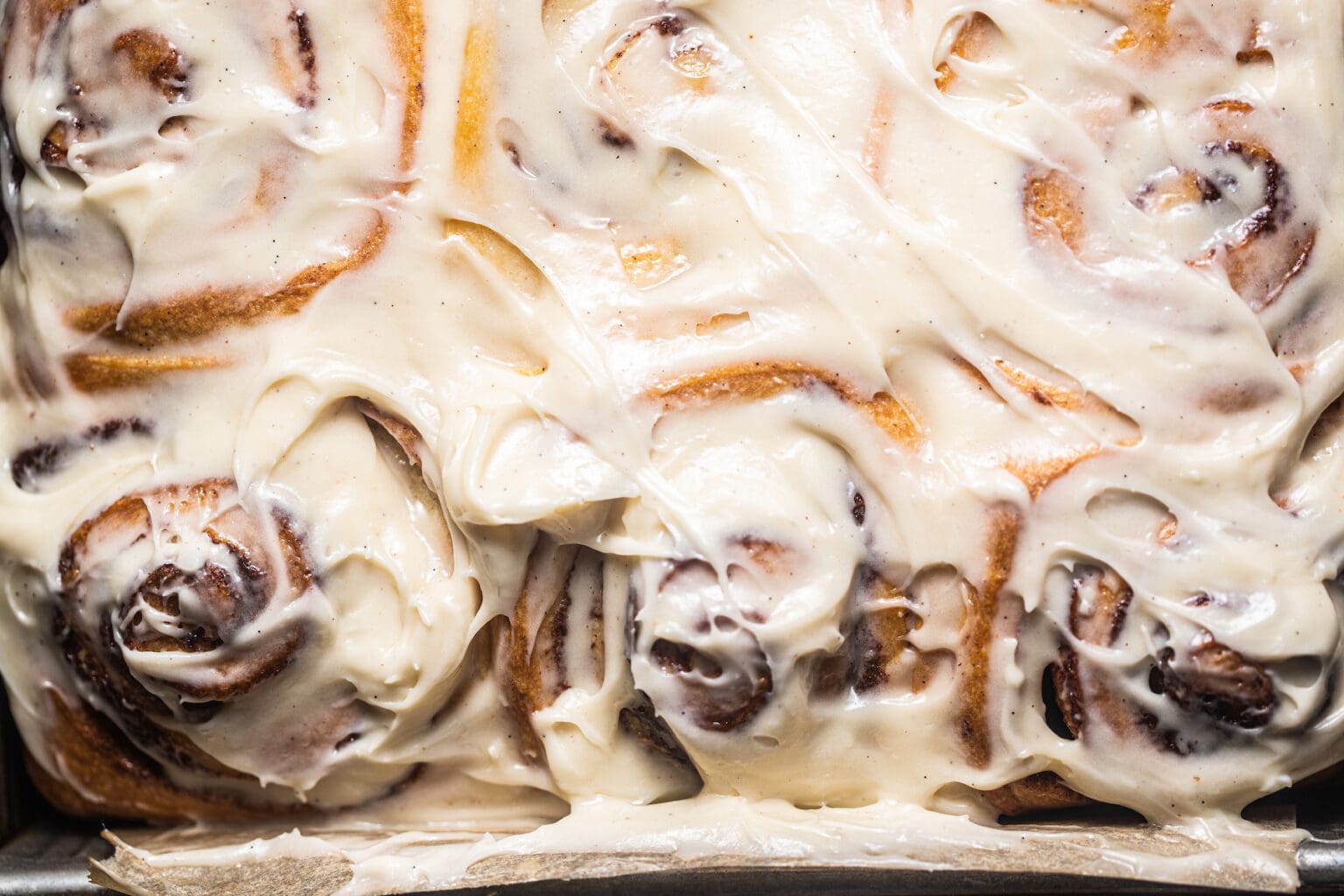 Close-up image of freshly baked cinnamon rolls generously covered in thick, creamy white frosting. The swirls of the rolls are clearly visible beneath the smooth icing, and the rolls have a golden-brown color, indicating they are perfectly baked.