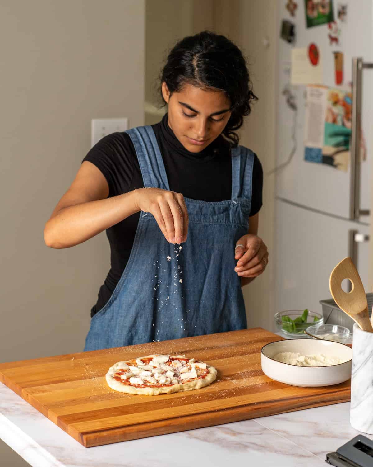 Woman sprinkling pizza dough with cheese on a cutting board.