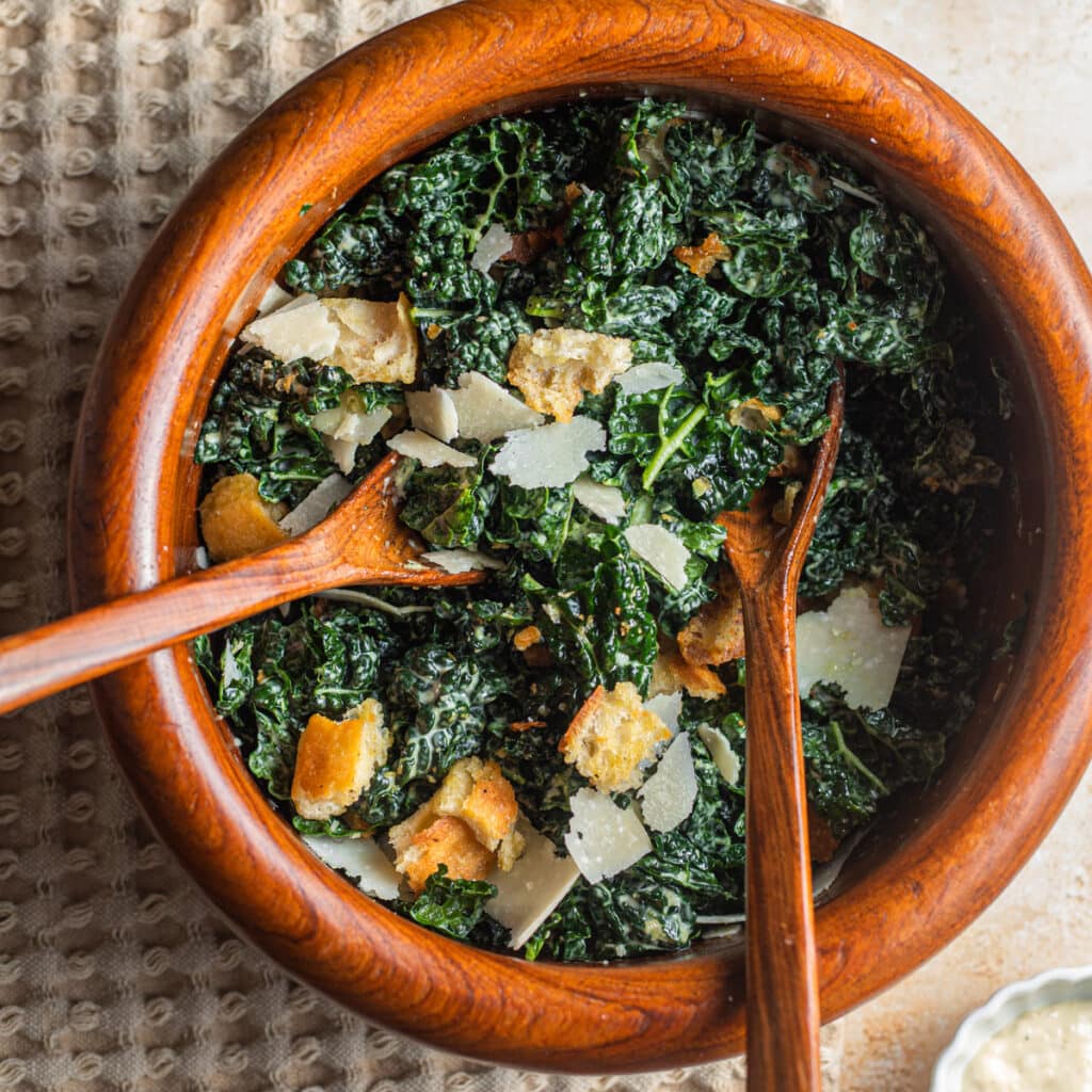 A large wooden bowl with wooden spoons filled with kale, croutons, and shaved cheese on a kitchen towel.