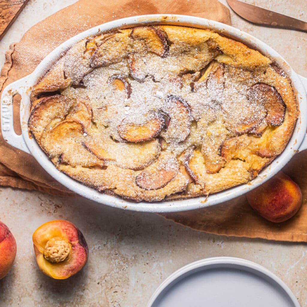 Casserole dish of peach clafoutis dusted with powdered sugar and peaches in the background