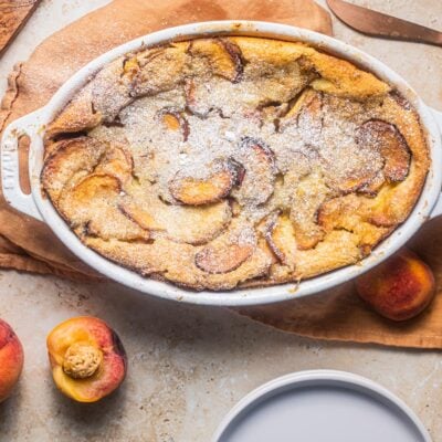Casserole dish of peach clafoutis dusted with powdered sugar and peaches in the background