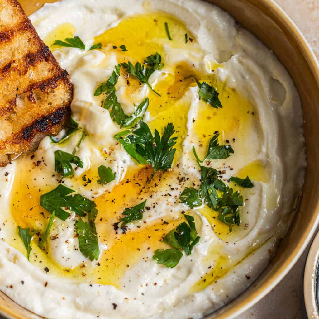 A bowl of whipped ricotta topped with olive oil, honey, black pepper, and fresh herbs with grilled bread in the background.