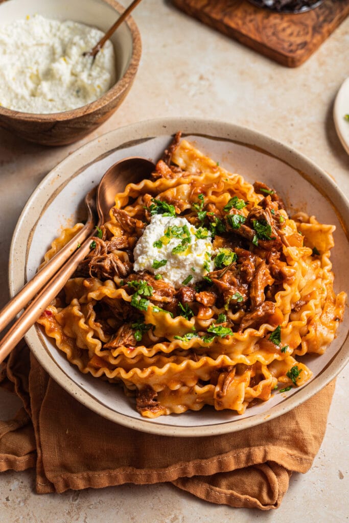 Bowl of lamb ragu and mafaldine pasta with a bowl of ricotta in the background.