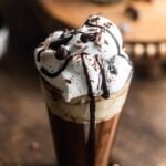 A glass of hot chocolate topped with whipped cream, chocolate flakes, and dripping chocolate syrup.