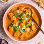 Bowl of butter paneer topped with cilantro and red onions with naan in the background.