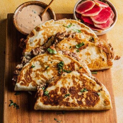 4 bean and cheese tacos arranged on a wooden cutting board with chipotle sauce and pickled radishes.