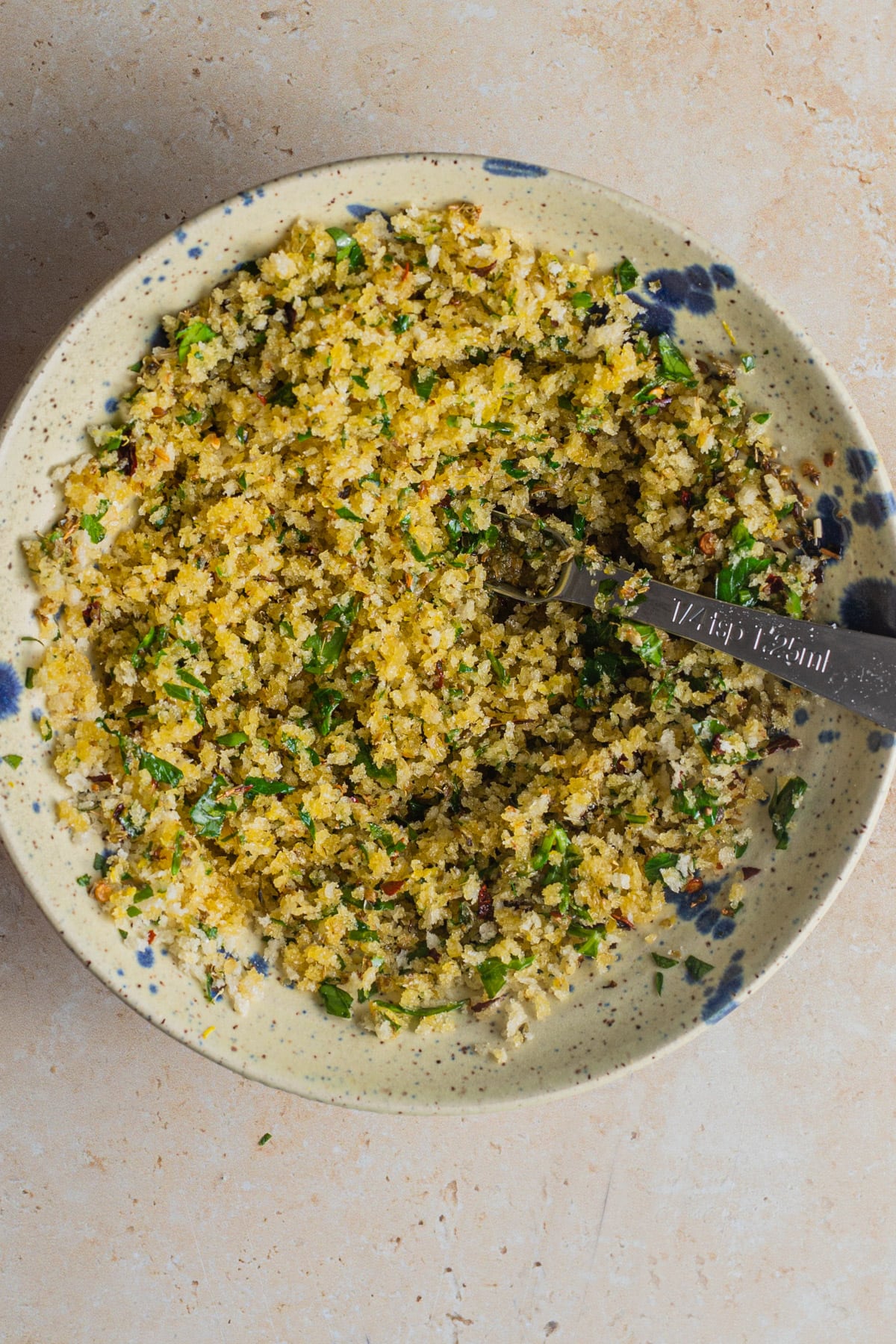 Crispy breadcrumbs tossed with parsley using a small measuring spoon in a speckled bowl.