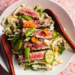 A bowl of shredded cabbage salad with slices of rare tuna garnished with cashews, orange segments, and two wooden spoons.