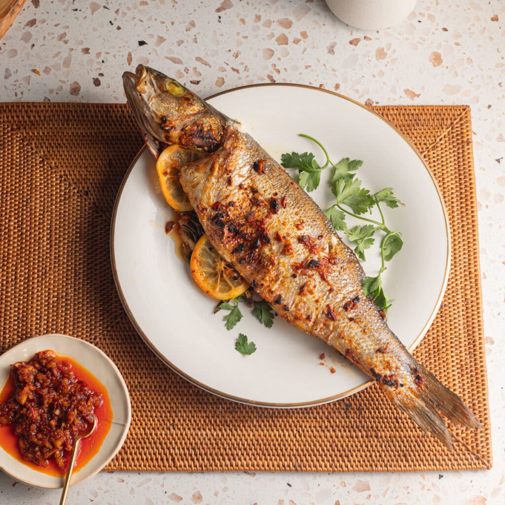 A whole-roasted branzino topped with a sauce and parsley on a plate set on a placemat.