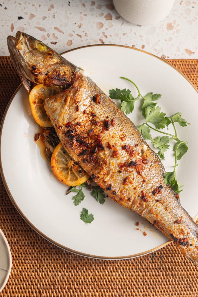 A whole-roasted branzino on a plate with sprigs of parsley and lemon.