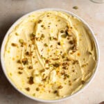 A bowl of shrikhand decorated with pistachios and saffron threads and a bowl of pistachios in the background.