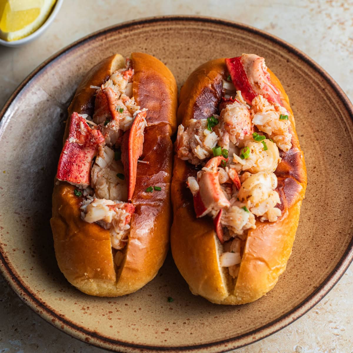 Two lobster rolls on a ceramic plate topped with chives and lemon wedges in the background.