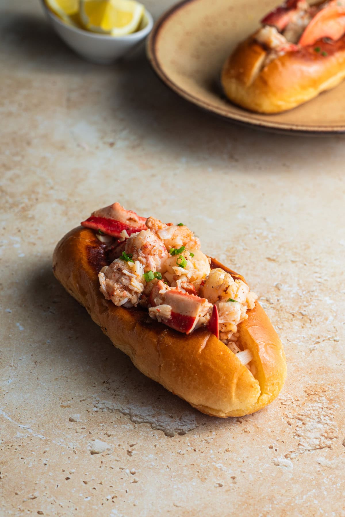 A lobster roll topped with chives with a lobster roll and lemon wedges in the background.