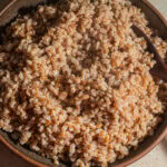Shallow ceramic bowl with cooked farro in it and a wooden spoon on a countertop.