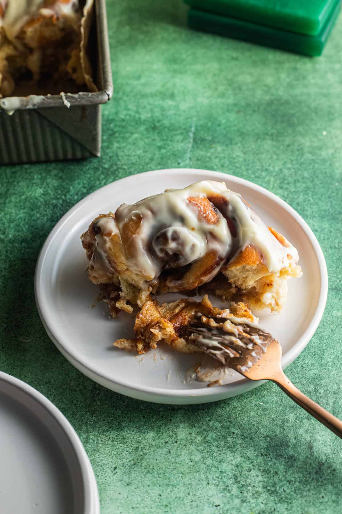 A half-eaten cinnamon roll on a plate with a fork and a metal baking pan in the background.