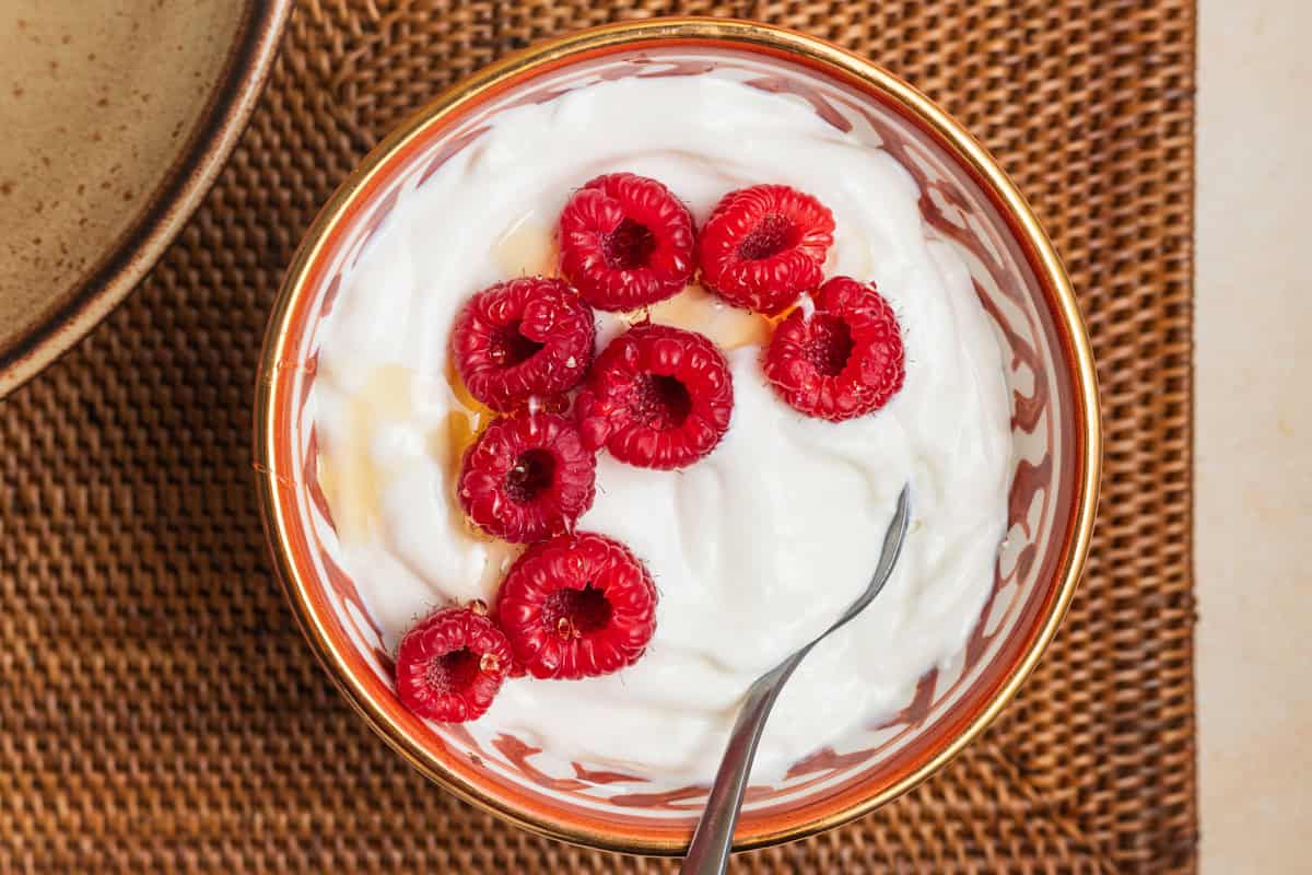 A bowl of Greek yogurt drizzled with honey and topped with raspberries set on a plate.
