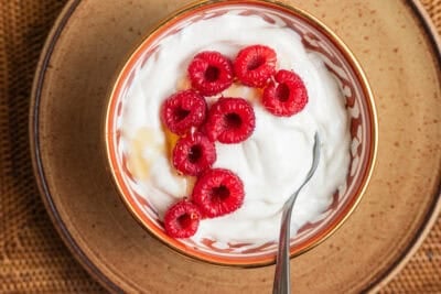 A bowl of Greek yogurt drizzled with honey and topped with raspberries set on a plate.