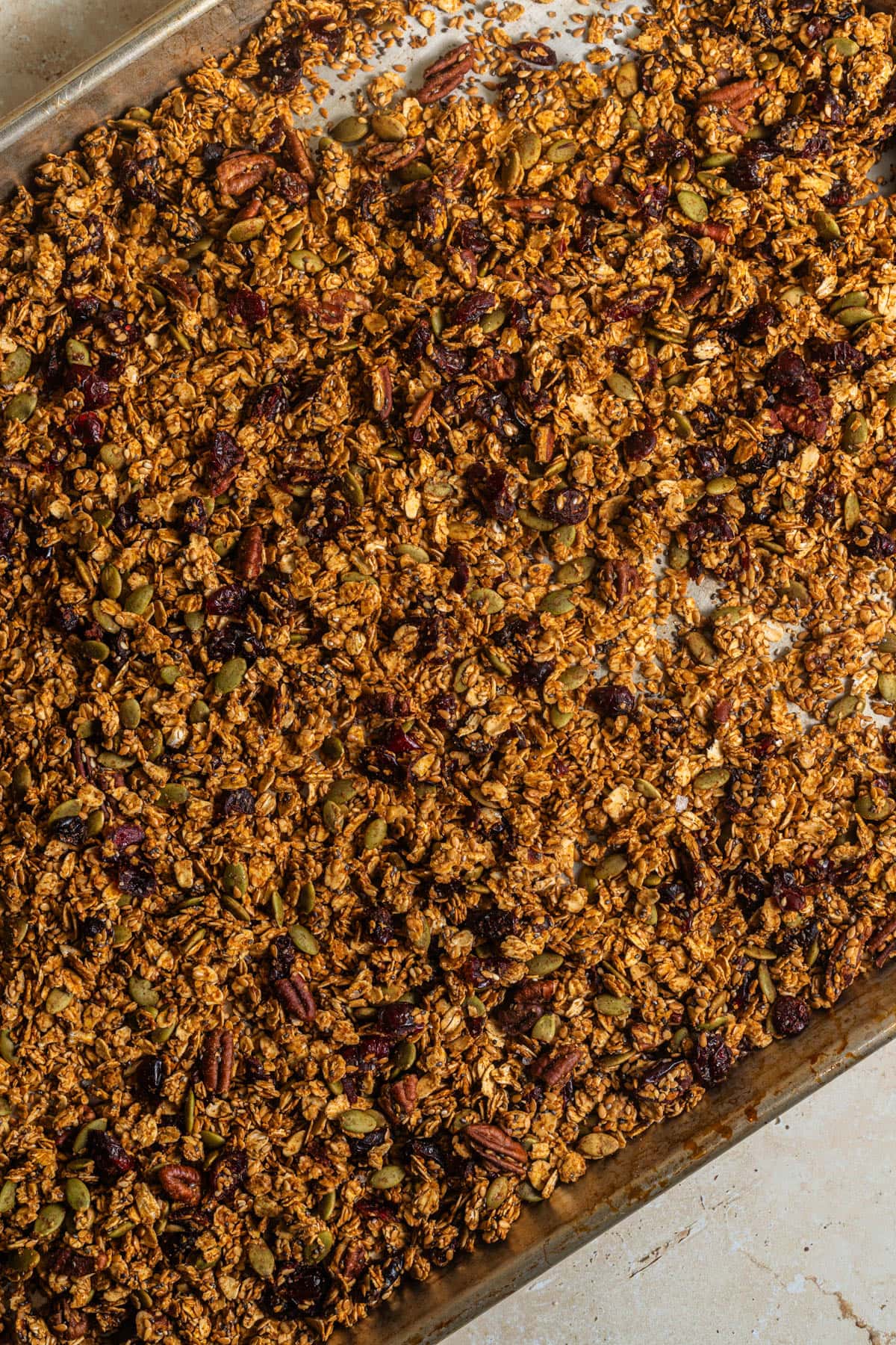 A stainless steel baking sheet with baked granola, cranberries, and pecans.