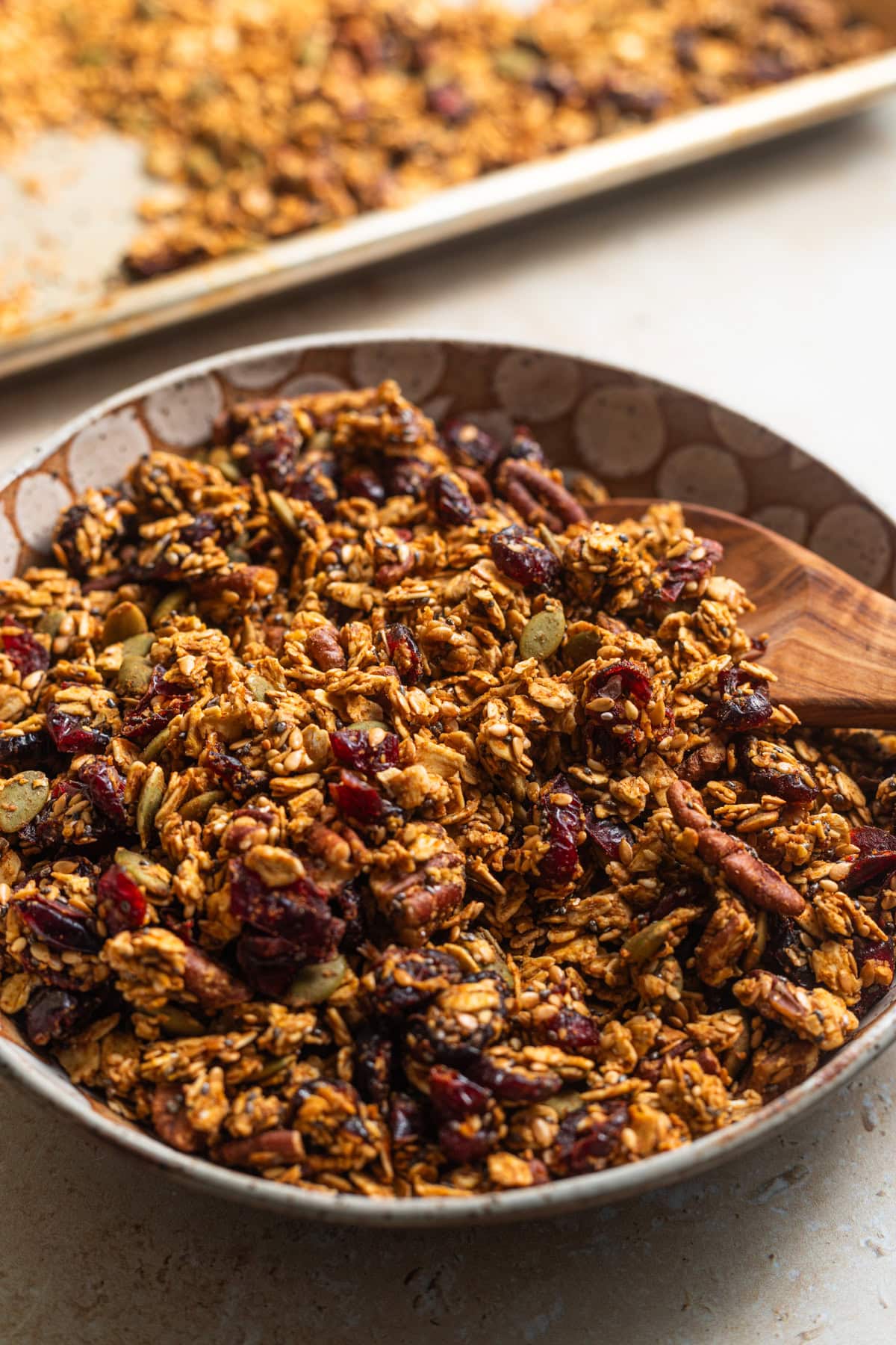 A bowl of granola with a wooden spoon and a baking sheet in the background.