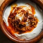 A wooden bowl with a poached egg sitting on a bed of yogurt doused with a chunky chili oil.