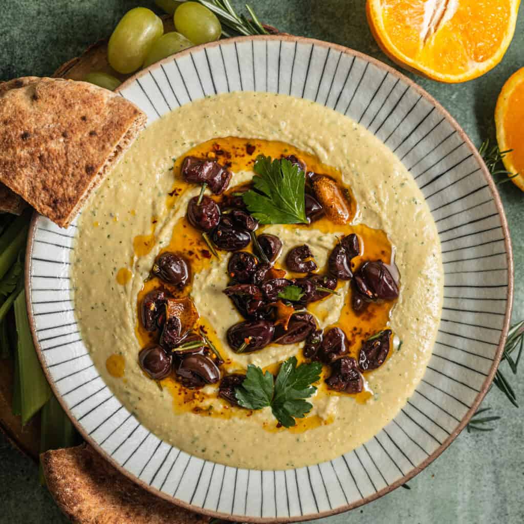 A bowl of white bean dip topped with olives and fresh herbs with fresh fruit in the background.